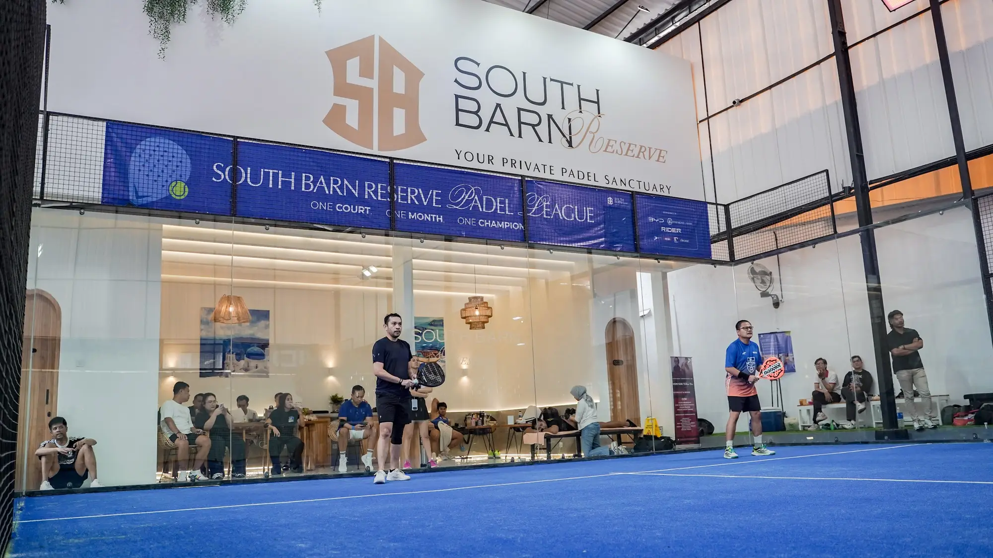Players on the South Barn Reserve court with the lounge visible behind the glass