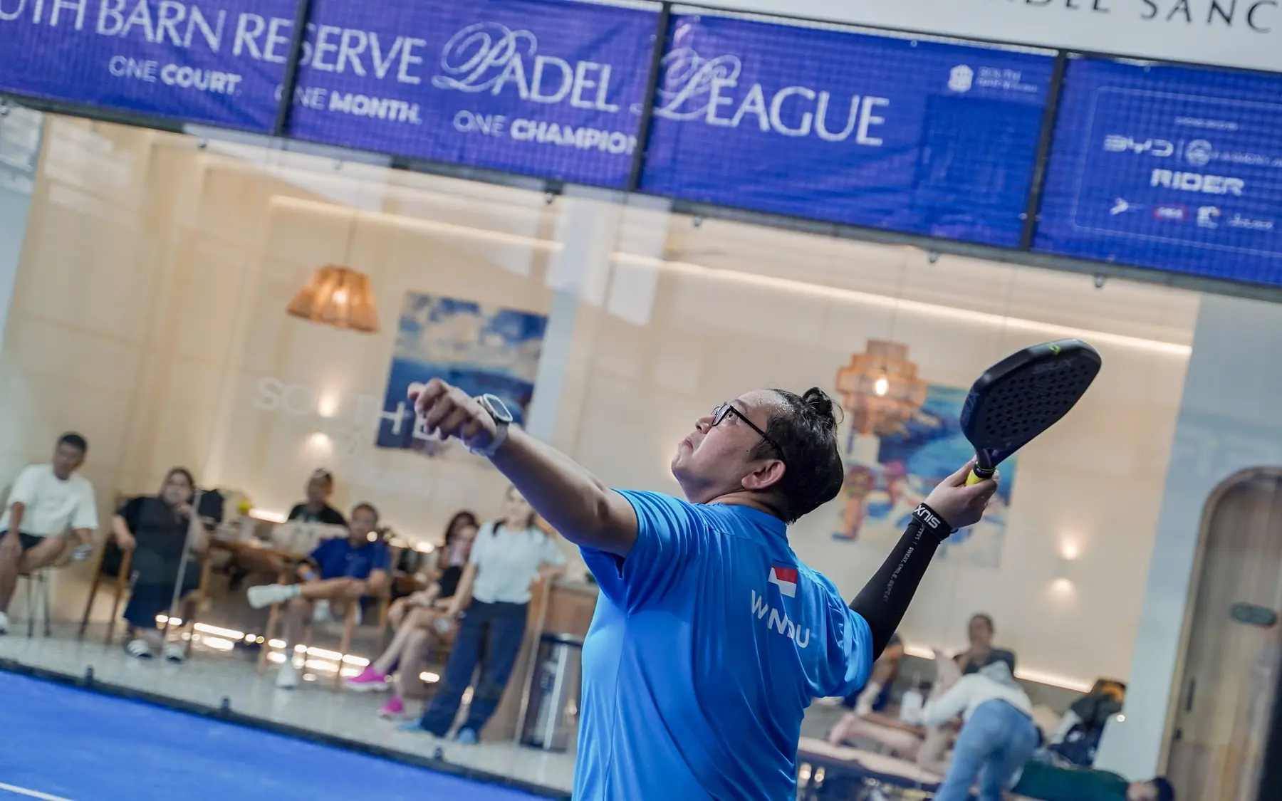A player serving while friends relax in the lounge behind the glass